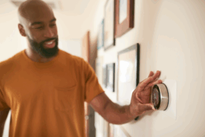 a man in an orange shirt turning the heat up on his home thermostat